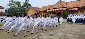 A group of people dressed in white uniforms are marching in formation in an open courtyard. In the background, there is a group of onlookers dressed in blue and white uniforms, standing in front of a building with tiled roofs and decorative banners. Some trees and other buildings are also visible in the background.