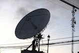 A large satellite dish is prominently positioned against the backdrop of a clear sky, intersected by numerous power lines. A nearby metal communication tower is visible, along with a lamp post. The scene is set in such a way that the structures are silhouetted due to the lighting conditions.
