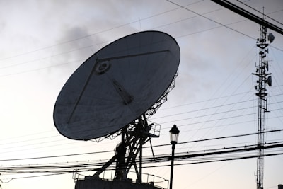 A large satellite dish is prominently positioned against the backdrop of a clear sky, intersected by numerous power lines. A nearby metal communication tower is visible, along with a lamp post. The scene is set in such a way that the structures are silhouetted due to the lighting conditions.