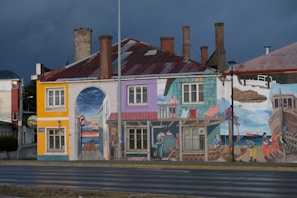 A building façade is painted with a vibrant mural depicting scenes of coastal life. The artwork features fishermen, boats, and a seaside town, with vivid colors and intricate details. The building's architecture is traditional with multiple chimneys and a rusty corrugated metal roof.