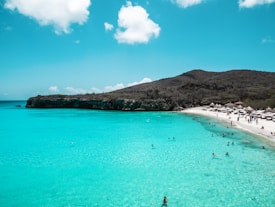 A serene beach with clear turquoise water, scattered with people swimming and relaxing. The shoreline curves along the edge with white sand, and the background features a hillside with sparse vegetation. A few beach huts and umbrellas are visible along the sand, providing shade to the beachgoers.