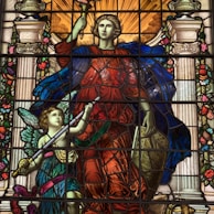 A colorful stained glass artwork depicting two figures. The central figure is regal, wearing a flowing red garment and a blue cloak, holding a torch and a shield. A smaller winged figure, possibly an angel, is beside them, holding a sword. The background includes ornate columns and floral decorations, with warm light emanating from above.