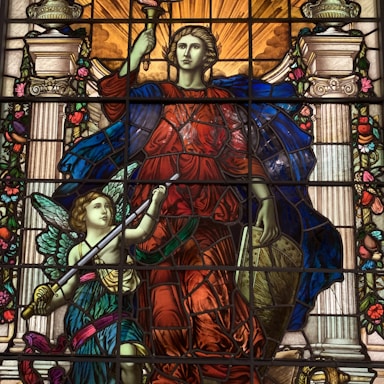 A colorful stained glass artwork depicting two figures. The central figure is regal, wearing a flowing red garment and a blue cloak, holding a torch and a shield. A smaller winged figure, possibly an angel, is beside them, holding a sword. The background includes ornate columns and floral decorations, with warm light emanating from above.