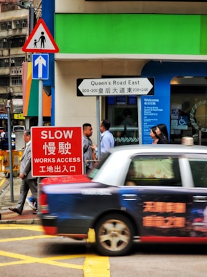 A bustling urban street scene showcases a taxi in motion, vibrant signage, and pedestrians walking near a colorful building facade. The upper portion features a mix of green and blue colors with informational signs indicating a road and access instructions.
