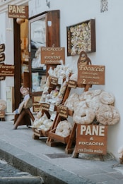 A rustic Italian bakery display on a street, featuring various types of bread and baked goods. Wooden signs with text in Italian advertise focaccia, panini, and other items made with sourdough. Large loaves and bags of bread are artfully arranged on shelves outside the shop, creating an inviting and traditional atmosphere.