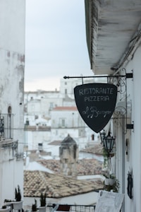 A charming street view in a historic town, featuring a rustic pizzeria sign hanging from a whitewashed building. In the background, there are more white-washed buildings with flat roofs and a dull sky, contributing to a nostalgic and serene atmosphere.
