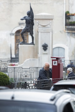 An elderly man is standing near a red parking meter in an outdoor setting. A statue of a soldier holding a flag is visible in the background. The area features architectural details such as a metal fence and a classical sculpture pedestal. There are cars parked in the foreground, and another individual is seen sitting in the background.