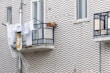 Clothes and a patterned cloth are drying on a metal balcony railing against a textured brick wall. A potted plant on the balcony adds greenery, and a plastic sheet is hanging below.