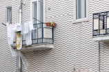 Clothes and a patterned cloth are drying on a metal balcony railing against a textured brick wall. A potted plant on the balcony adds greenery, and a plastic sheet is hanging below.