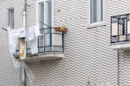 Clothes and a patterned cloth are drying on a metal balcony railing against a textured brick wall. A potted plant on the balcony adds greenery, and a plastic sheet is hanging below.