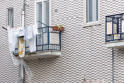 Clothes and a patterned cloth are drying on a metal balcony railing against a textured brick wall. A potted plant on the balcony adds greenery, and a plastic sheet is hanging below.
