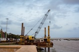 A large crane is positioned on a platform by the riverbank. It appears to be part of ongoing construction or maintenance work along the river. The crane is surrounded by other machinery and a few personnel can be seen nearby, indicating industrial activity. In the background, there is a bridge spanning the river, and the sky is overcast with scattered clouds.