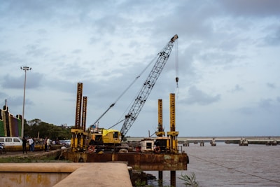 A large crane is positioned on a platform by the riverbank. It appears to be part of ongoing construction or maintenance work along the river. The crane is surrounded by other machinery and a few personnel can be seen nearby, indicating industrial activity. In the background, there is a bridge spanning the river, and the sky is overcast with scattered clouds.