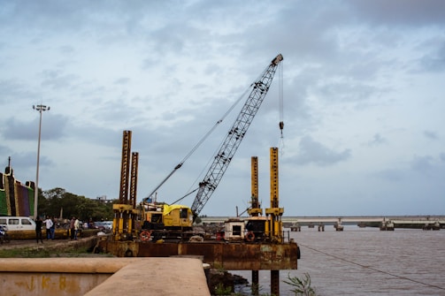 A large crane is positioned on a platform by the riverbank. It appears to be part of ongoing construction or maintenance work along the river. The crane is surrounded by other machinery and a few personnel can be seen nearby, indicating industrial activity. In the background, there is a bridge spanning the river, and the sky is overcast with scattered clouds.