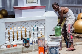 A person is standing next to a small wall, which appears to be freshly painted white. There are several large paint containers, a green bottle, and some paintbrushes on the ground. The person, wearing casual clothing, has paint on their skin and clothes, suggesting they are in the process of painting. Large golden spherical objects can be seen in the background.