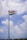 A tall flagpole with the Indian national flag waving against a partly cloudy blue sky. There are a few people and a stone wall at the base of the flagpole, possibly part of a fort or a similar structure.