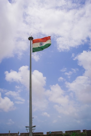 A tall flagpole with the Indian national flag waving against a partly cloudy blue sky. There are a few people and a stone wall at the base of the flagpole, possibly part of a fort or a similar structure.
