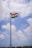 A tall flagpole with the Indian national flag waving against a partly cloudy blue sky. There are a few people and a stone wall at the base of the flagpole, possibly part of a fort or a similar structure.