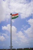 A tall flagpole with the Indian national flag waving against a partly cloudy blue sky. There are a few people and a stone wall at the base of the flagpole, possibly part of a fort or a similar structure.