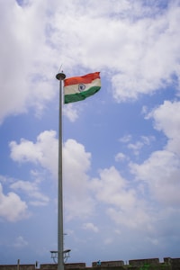 A tall flagpole with the Indian national flag waving against a partly cloudy blue sky. There are a few people and a stone wall at the base of the flagpole, possibly part of a fort or a similar structure.