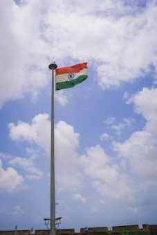 A tall flagpole with the Indian national flag waving against a partly cloudy blue sky. There are a few people and a stone wall at the base of the flagpole, possibly part of a fort or a similar structure.