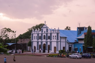 A large, white and blue church building with a triangular roof, ornate architectural details, and a cross on top. The church is situated in an open space with a few trees in the background. Several people are walking around the vicinity, and there are cars and motorcycles parked nearby. The sky has a mix of light and darker clouds, indicating an early evening or late afternoon setting.