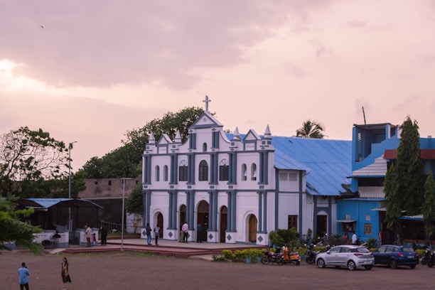 A large, white and blue church building with a triangular roof, ornate architectural details, and a cross on top. The church is situated in an open space with a few trees in the background. Several people are walking around the vicinity, and there are cars and motorcycles parked nearby. The sky has a mix of light and darker clouds, indicating an early evening or late afternoon setting.