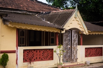 An old house with a tiled sloping roof, featuring intricate white metal latticework on the front door. The building is primarily painted yellow with rustic red decorative accents and railings on the veranda. Small plants in pots are placed near the entrance, adding a touch of greenery.