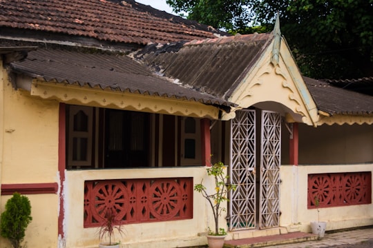 An old house with a tiled sloping roof, featuring intricate white metal latticework on the front door. The building is primarily painted yellow with rustic red decorative accents and railings on the veranda. Small plants in pots are placed near the entrance, adding a touch of greenery.