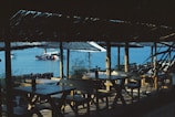 A seaside dining area with wooden tables and benches under thatched roofs. In the distance, a boat is anchored on a calm blue sea. The scene includes natural light casting shadows under the roofs, creating a serene and tranquil atmosphere.