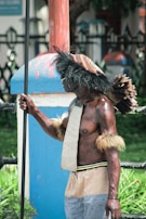 A person wearing traditional attire adorned with feathers and holding a spear. The costume includes a headdress made of woven fibers and feathers, arm bands with fur, and a loincloth. The person stands near a blue and red post, surrounded by greenery.
