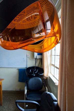 An interior space of a hair salon featuring an orange hair dryer hood prominently in the foreground. Below it is a black salon chair and a washing station. Natural light filters through large windows with beige curtains, casting a warm ambiance. The room has a minimalist decor, with a whiteboard and a small table visible in the background.