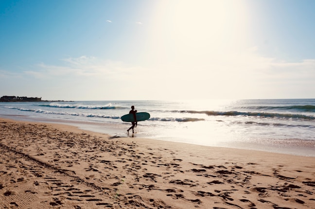 A surfer carrying a surfboard walks along the beach towards the ocean. The sun is shining brightly, casting a warm glow over the scene. The gentle waves are lapping at the shore, and footprints are visible in the sand.