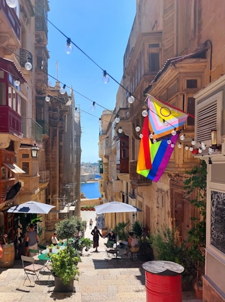A narrow cobblestone street lined with old buildings featuring wooden balconies and shutters. A vibrant pride flag is prominently displayed, and strings of lights are hung above the street. A few people are sitting at tables with umbrellas in an outdoor cafe, enjoying sunny weather. In the background, there is a view of a harbor with blue water and distant buildings.