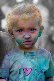 A young child with colorful powder on their face and clothes, likely at a festival or event involving color. The child has blonde curly hair and blue eyes, wearing a t-shirt with a cat design holding a drink, accompanied by text. The background is blurred with muted people present.