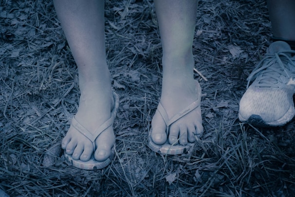 Feet wearing flip-flops stand on dried grass and leaves. Another foot in a running shoe is partially visible to the side.