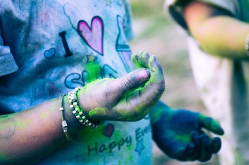 A person wearing a t-shirt with colorful designs, featuring text and symbols. Their hands are covered in vibrant green and blue powder, suggesting participation in a festival or color event. They wear bracelets and the background is blurred, with another person partially visible.