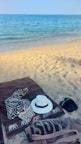 A sandy beach with clear blue ocean water in the background. On the sand lies a brown towel with a zebra-patterned cloth and a white hat. Nearby, there are black flip-flops and a beige tote bag.