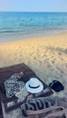A sandy beach with clear blue ocean water in the background. On the sand lies a brown towel with a zebra-patterned cloth and a white hat. Nearby, there are black flip-flops and a beige tote bag.