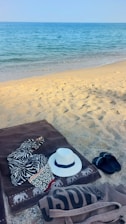 A sandy beach with clear blue ocean water in the background. On the sand lies a brown towel with a zebra-patterned cloth and a white hat. Nearby, there are black flip-flops and a beige tote bag.