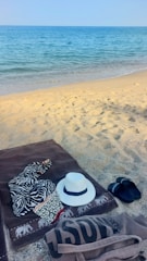 Close-up of a beachcraft tote bag resting on a sandy towel beside sunglasses and a sunhat.
