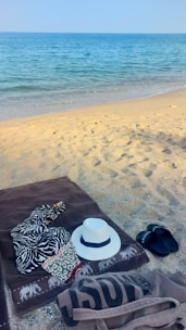 Close-up of a beachcraft tote bag resting on a sandy towel beside sunglasses and a sunhat.