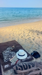 A sandy beach with clear blue ocean water in the background. On the sand lies a brown towel with a zebra-patterned cloth and a white hat. Nearby, there are black flip-flops and a beige tote bag.
