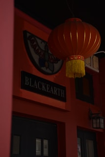 A dark red lantern with gold accents hangs from the ceiling in front of a sign that reads 'BLACKEARTH'. The background features orange walls with a stained glass or painted emblem depicting a butterfly shape and a black-framed door.