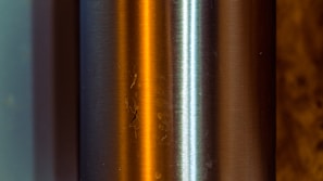 A close-up of a metallic cylindrical object with a brushed finish. The surface reflects warm orange and cool blue light, creating an interesting contrast. The two main strips of color highlight the curvature and texture of the metal, while small imperfections and scratches are visible.