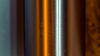 A close-up of a metallic cylindrical object with a brushed finish. The surface reflects warm orange and cool blue light, creating an interesting contrast. The two main strips of color highlight the curvature and texture of the metal, while small imperfections and scratches are visible.