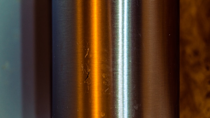 A close-up of a metallic cylindrical object with a brushed finish. The surface reflects warm orange and cool blue light, creating an interesting contrast. The two main strips of color highlight the curvature and texture of the metal, while small imperfections and scratches are visible.