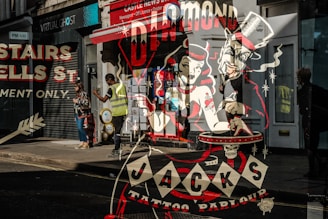 A vibrant street scene with reflections in a shop window showcasing vintage style artwork for 'Jack's Tattoo Parlour'. The artwork features retro characters and classic tattoo imagery, blending with real-life pedestrians, including a person in a yellow vest and a woman in casual attire. The shop signs and bright colors create an urban atmosphere.