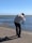 A person stands on the edge of a body of water, adjusting a camera on a tripod. The scene depicts a serene coastal environment with gentle waves and a group of flamingos in the background. The person is dressed in a light vest and dark pants, capturing the moment against a clear blue sky.