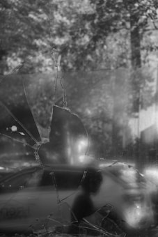 A cracked window with a large fracture dominates the foreground, against a blurred backdrop of a vehicle and a person's silhouette. The scene is captured in black and white, adding a somber, reflective quality to the image.